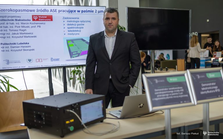 profesor stoi, za sobą ma ekran z prezentacją, przed nim na stole stoi urządzenie